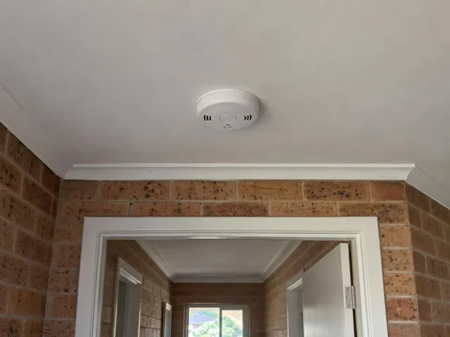 White smoke alarm on a hallway ceiling above exposed brick walls and a doorway in Lake Munmorah